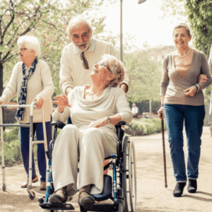 A group of 6 smiling older adults, many with mobility aids, strolls through a park together.