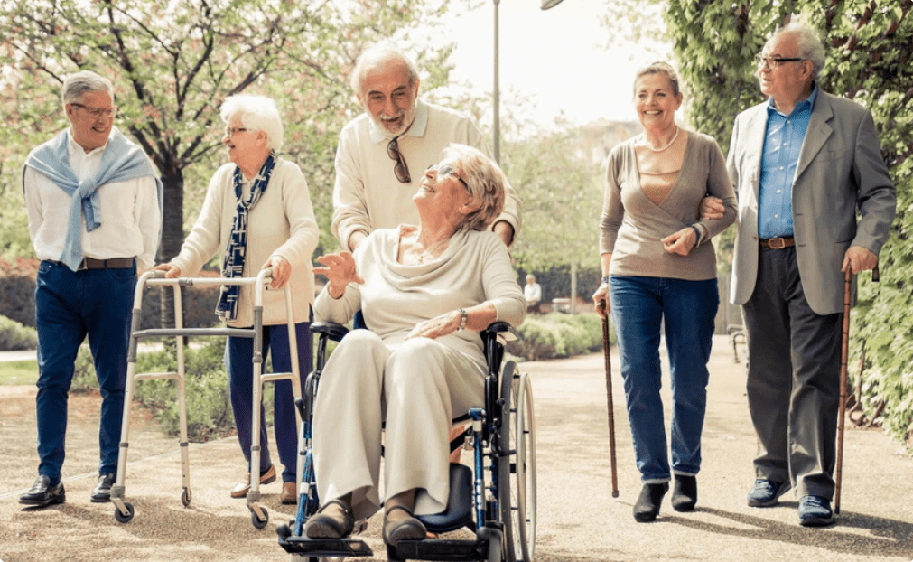 A group of 6 smiling older adults, many with mobility aids, strolls through a park together.