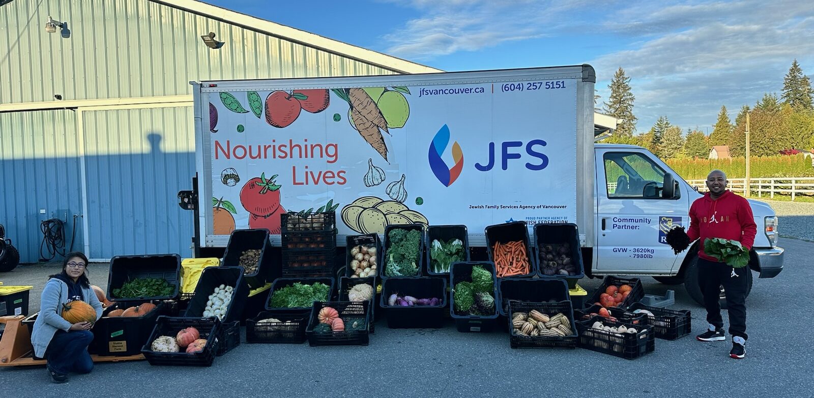 Plastic bins filled with a variety of fresh produce is displayed in front of the JFS truck. Two people pose with the bins. In the background are a barn and blue sky.