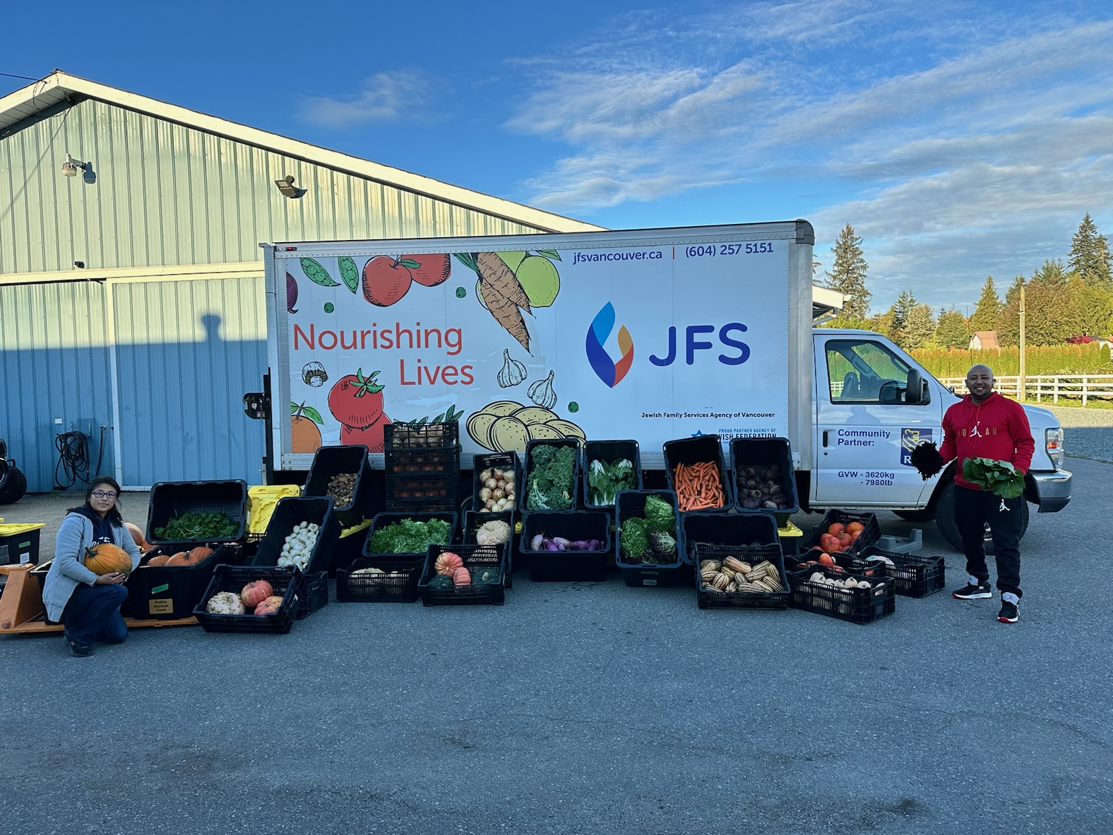 Plastic bins filled with a variety of fresh produce is displayed in front of the JFS truck. Two people pose with the bins. In the background are a barn and blue sky.