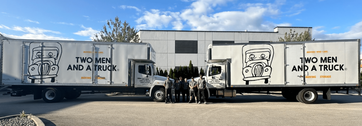 A small group of people post in front of two large moving trucks with the Two Men and a Truck logo. A building and blue sky are in the background.